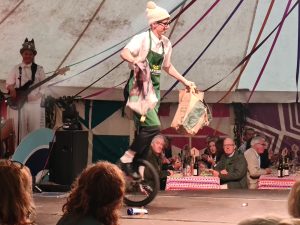 The clown riding a unicycle holding two bags